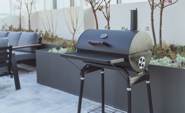 A black, kettle-style charcoal grill standing on a patio next to gray-upholstered outdoor furniture and elevated garden planters.