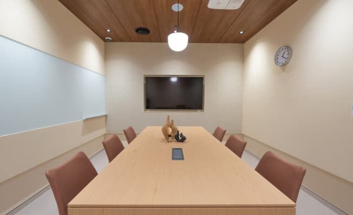 Formal meeting room with a light wood table, comfortable brown chairs, a mounted television screen, and a large whiteboard.