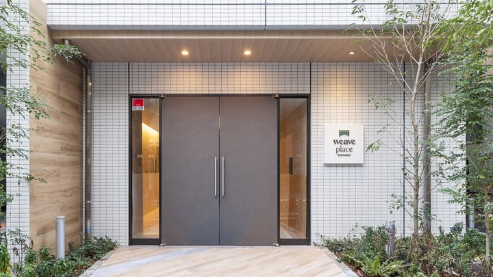 Exterior view of the main entrance to Weave Place Ryogoku, featuring modern, dark gray double doors, white tiled walls, and small trees and landscaping on either side.