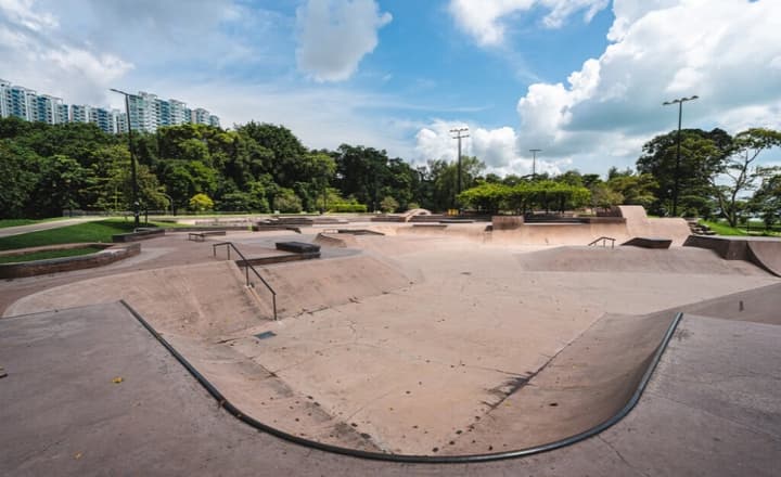 Wide view of a large, empty concrete skatepark with various ramps, bowls, and rails, backed by lush green trees and high-rise apartments.