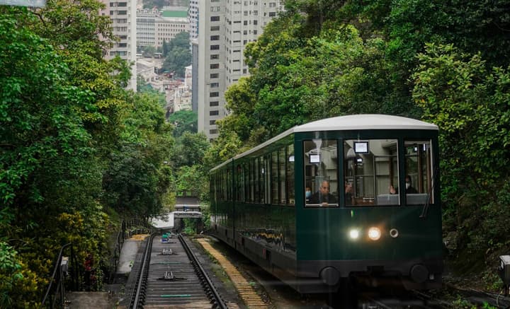 The green Peak Tram funicular ascending a steep track through dense trees and foliage, with tall Hong Kong residential buildings visible in the background.