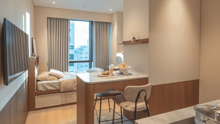 Bright, open-concept urban studio suite featuring a wooden kitchen bar/dining table with stools separating the cooking area from the cozy bedroom and large window.