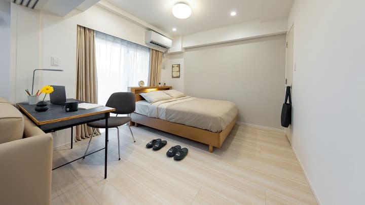 Focused view of the bedroom area with a wooden bed frame, a black modern desk and chair, and large window with light curtains.