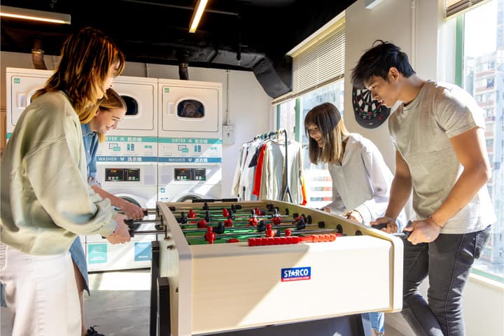 Four young adults playing foosball and laughing in a bright communal laundry room that features several washing machines and dryers.