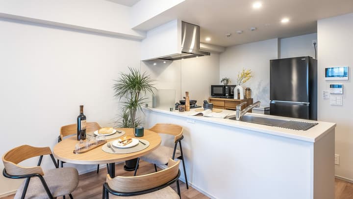Contemporary kitchen with a light wood dining table and four chairs next to a white kitchen counter, showing a dark refrigerator and a potted plant.