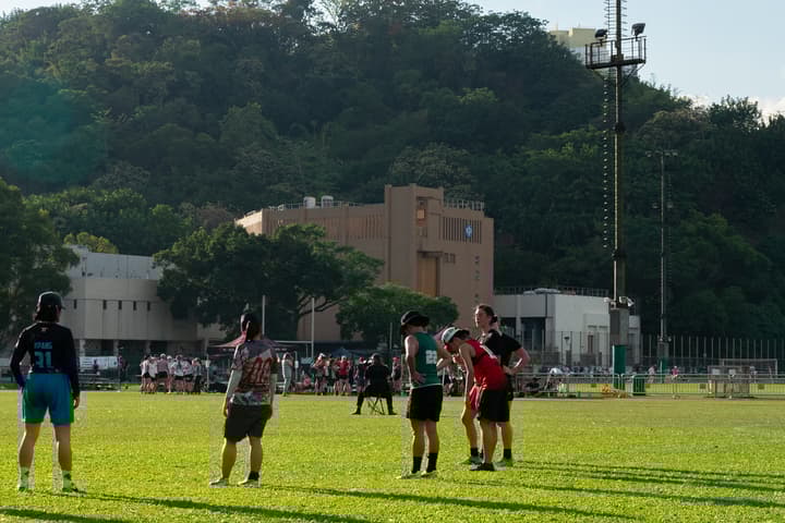 A group of Ultimate Frisbee players resting or waiting on a grassy outdoor sports field, with a large, forested mountain and a university-style building in the background.