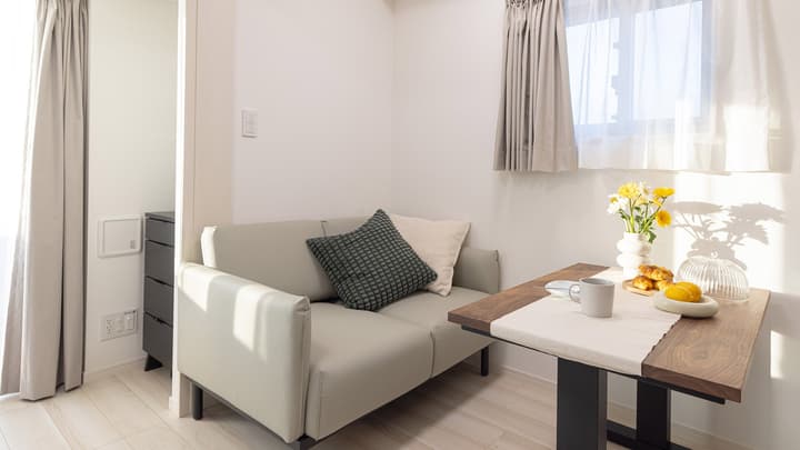 Compact living and dining area with a small, light gray two-seater sofa and a dark wood table set with a mug and pastries, bathed in sunlight.