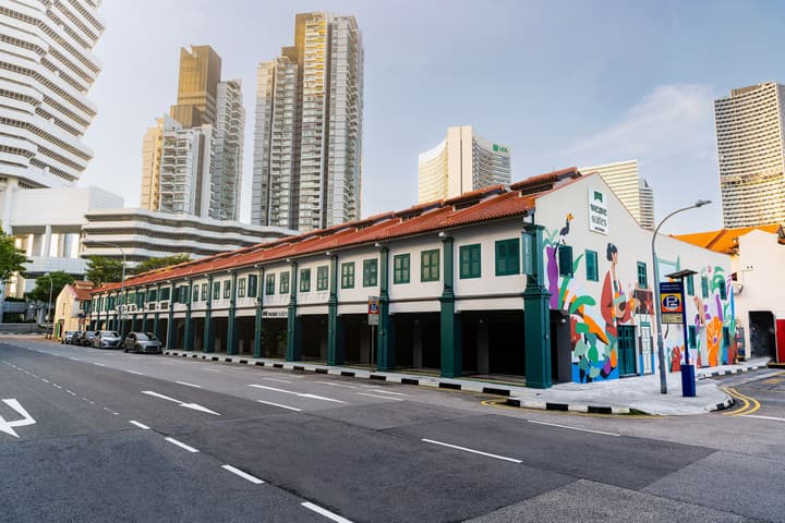 Wide exterior view of a row of historic shophouses with a terracotta roof and teal accents, featuring a large modern mural on one end, set against a backdrop of the Singapore city skyline.