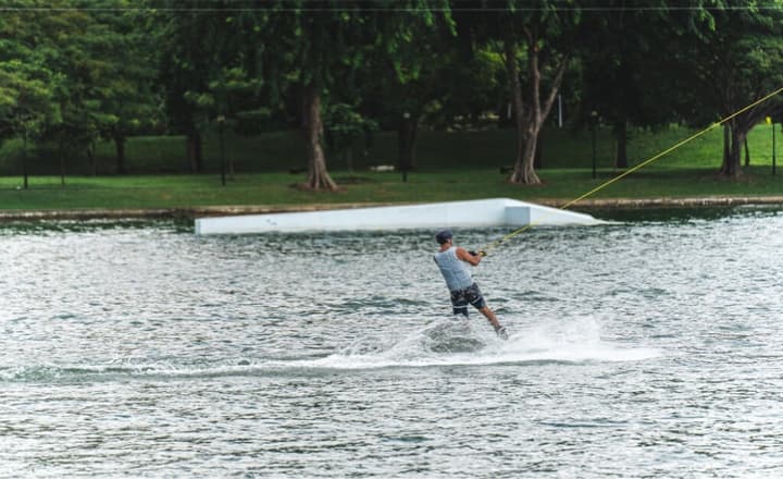 A man wakeboarding across a lake, carving a spray of water behind him, with a grassy park and trees visible in the background.