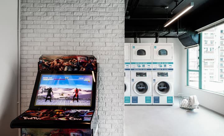A fun, shared laundry room with white stackable washers and dryers visible through a doorway, and an arcade fighting game machine set against a white exposed brick wall.