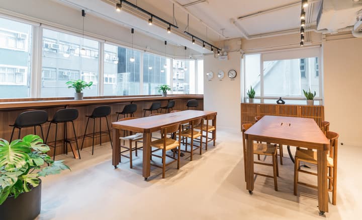 Modern, open co-working space with wooden long tables for collaboration, matching bar-height seating against a window counter, and world clocks on the wall, overlooking the city skyline.