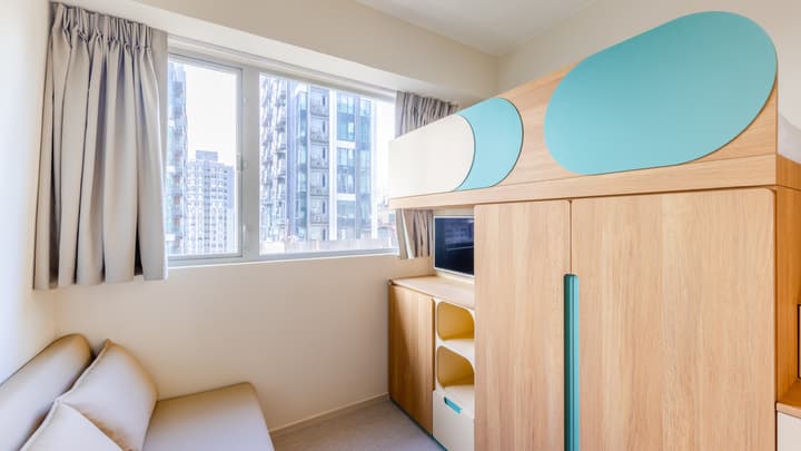 View of a compact studio bedroom featuring a wooden bed/storage unit with rounded mint-green and white panels, a window with curtains, and a small sofa below.