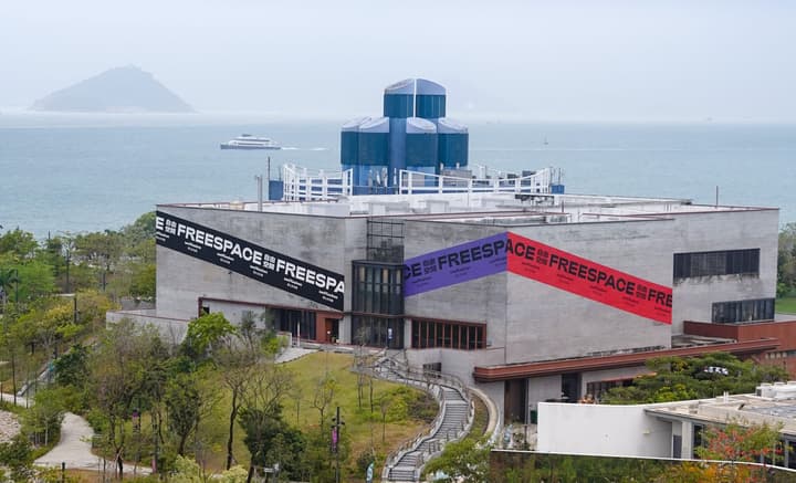 Wide exterior view of a modern, low-rise concrete building with a distinct blue, turret-like structure on its roof. The facade is marked with large "FREESPACE" banners in black, purple, and red, set against a background of the sea and distant islands.