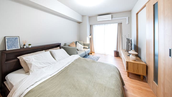 Full view of the studio apartment, showing the double bed in the foreground, a gray sofa, TV on a wooden stand, and a glass door leading to the balcony in Monzennakacho.