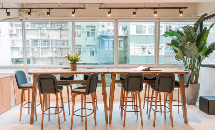 A bright communal dining or work area featuring two long wooden high tables surrounded by black cushioned bar stools, set in front of a wide window overlooking light green and white city buildings.