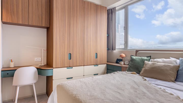 View of a queen-sized bed next to a large window with a harbour view, showing a floor-to-ceiling built-in wardrobe and a small integrated study desk with a chair.