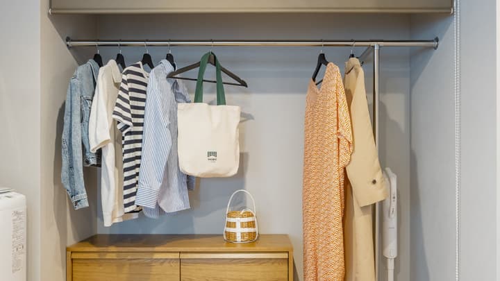 Open wardrobe area with a clothes rail and a two-drawer wooden dresser underneath, displaying various casual clothes, a tote bag, and a small woven basket.