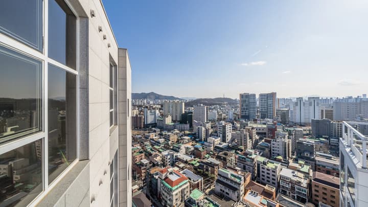 Bright, high-angle view of a densely packed city on a sunny day, showing a mix of low-rise and high-rise buildings and distant hills.