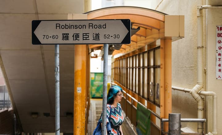 Street-level view of a 'Robinson Road' sign in English and Chinese, with a person walking on a covered, elevated pedestrian walkway in the background.