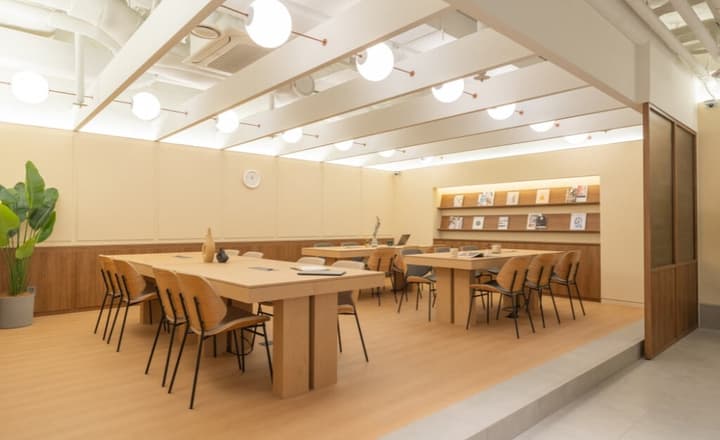 Bright, minimalist co-working or meeting room with light wood furniture, multiple large tables, and decorative shelving.