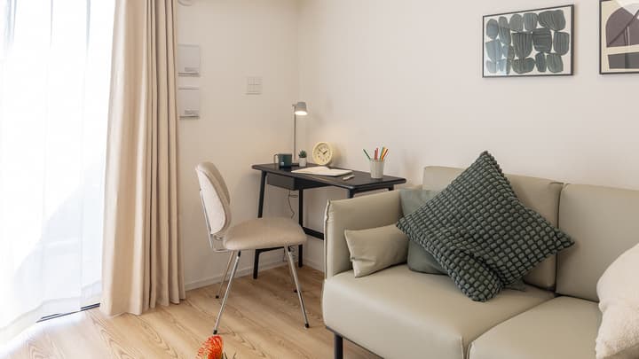 A view of the living room with a light beige sofa, a compact black desk and chair for working, and sheer curtains covering the large sliding door to the balcony.