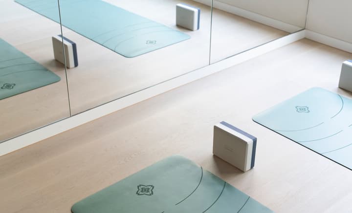 A bright, clean yoga or fitness studio floor with three light blue yoga mats and white blocks placed on light wood flooring, reflecting in a full-length mirrored wall.