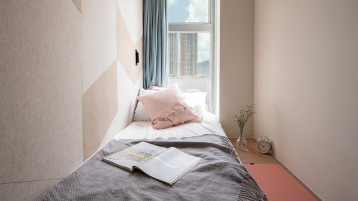 A close-up of a single bed with white bedding and a pink decorative pillow. An open book and a gray throw blanket are on the bed. The wall features a modern, geometric pattern of light beige and cream tones, while a tall window shows a city skyline and has teal curtains.