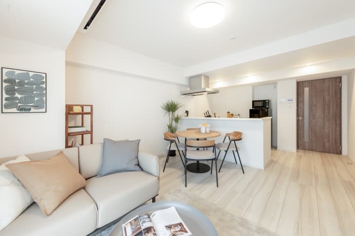 View of a bright, minimalist apartment living area from behind the sofa, showing the dining table with modern chairs and the white kitchen counter in the background.