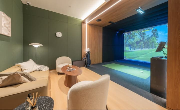 Indoor golf simulator screen next to a small lounge area with a tan couch, accent chairs, and a dark green paneled wall.