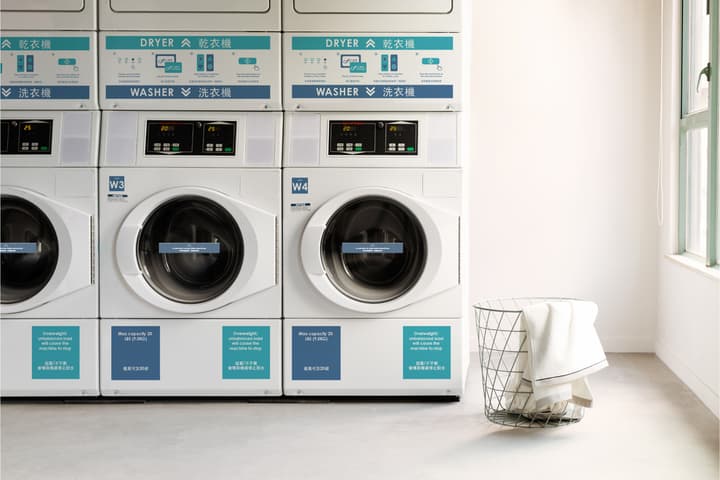 A brightly lit communal laundry room with a row of clean, stacked white washer and dryer units, next to a wire laundry basket overflowing with white towels.