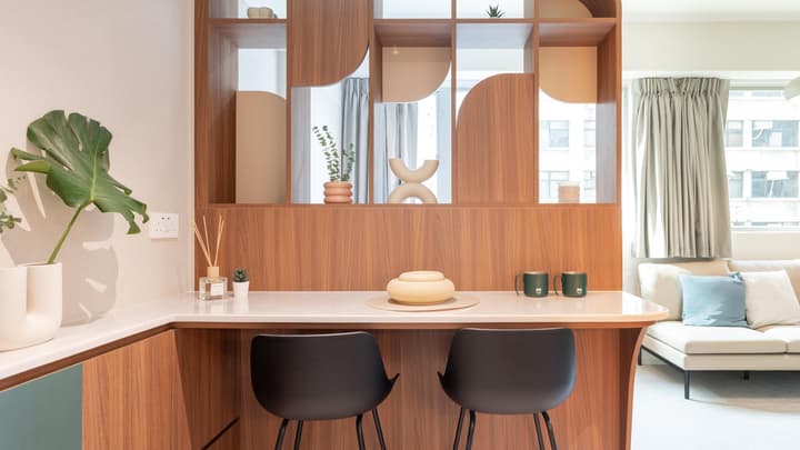 Close-up of a built-in dining bar with two black stools, a white countertop, and a wooden room divider/storage shelf with geometric cutouts separating the dining and living areas.