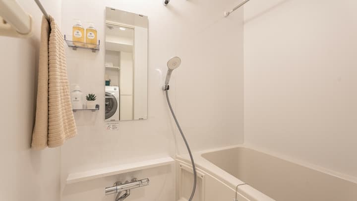 All-white unit bathroom with a deep bathtub, a hand-held shower head, a small mirror, a shelf for toiletries, and a tan hand towel hanging on a bar.