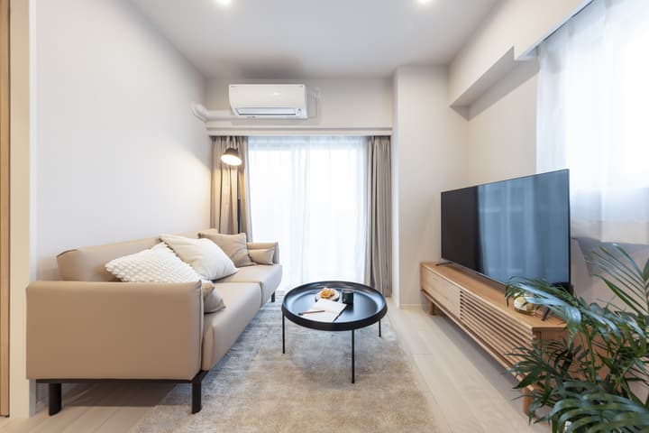 Bright living room featuring a tan sofa, a round black coffee table, a large flat-screen TV on a slatted light wood console, and sheer curtains covering a sliding glass door.