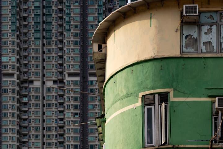 Extreme contrast between an older, weathered yellow and green cylindrical building and a towering, dense modern residential skyscraper in the background.