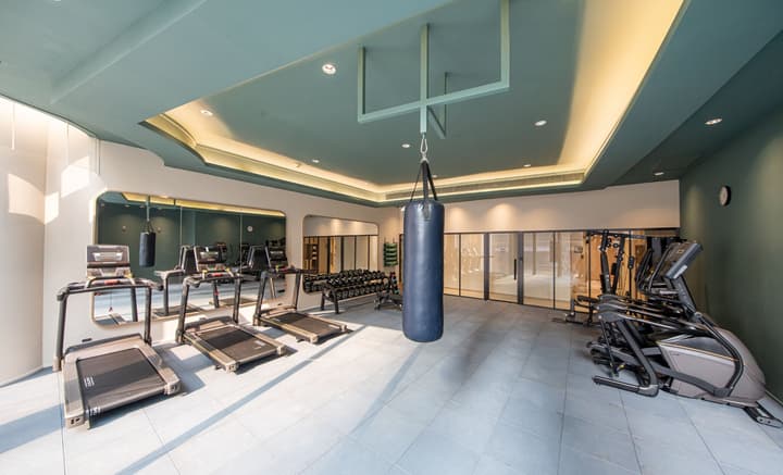 Interior view of a gym or fitness room with a punching bag hanging from the ceiling. Equipment includes several treadmills and a rack of free weights, with a wall of mirrors and a glass partition separating areas. The ceiling is painted a pale teal color.