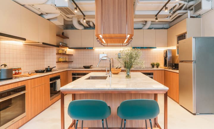 Spacious, modern communal kitchen featuring wood cabinetry, light pink tiled backsplash, a central marble island with a sink and two turquoise bar stools, and stainless steel full-size appliances.