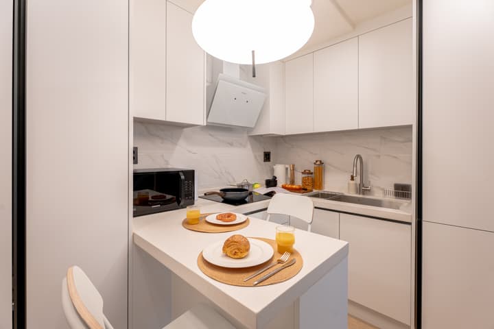 Bright, all-white compact kitchen with a marble-effect backsplash, an electric cooktop, a microwave, and a small counter bar used as a dining space for two.