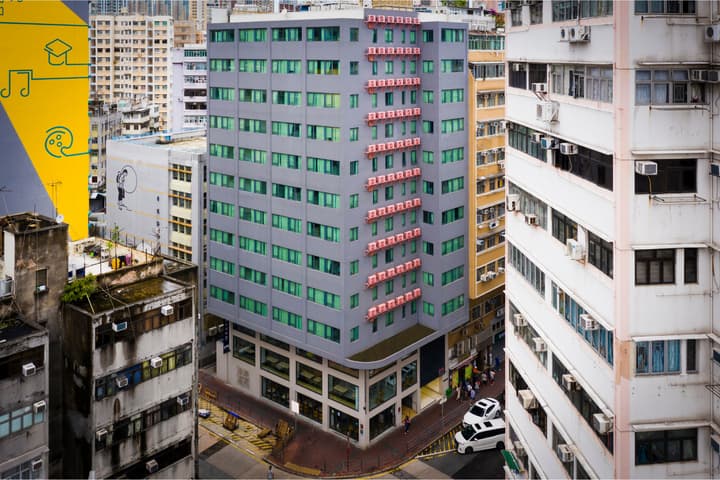 An aerial view looking down onto a cluster of contrasting Hong Kong buildings, including a prominent modern gray structure with green windows, surrounded by older residential blocks.