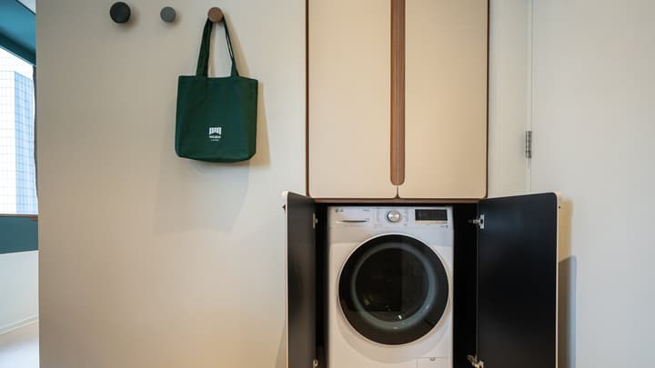 A front-loading washer or dryer neatly housed inside a cabinet with the doors open. Above the machine is another small, closed cabinet. To the left, a green tote bag hangs on a wooden wall hook.