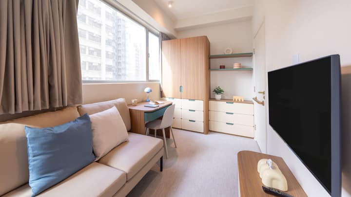 Bright living area of a premier studio with a beige sofa, a small desk, and a tall, wood-grain wardrobe/storage unit next to a large window.