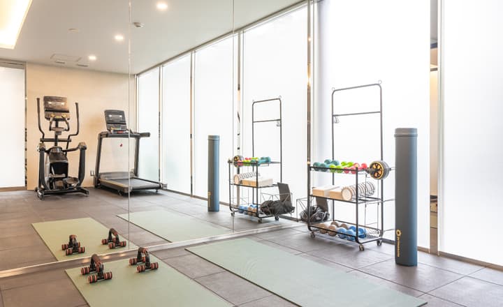 Building amenity image of a modern, bright communal gym with a treadmill, elliptical machine, floor mats, push-up bars, weights, and a mirrored wall.