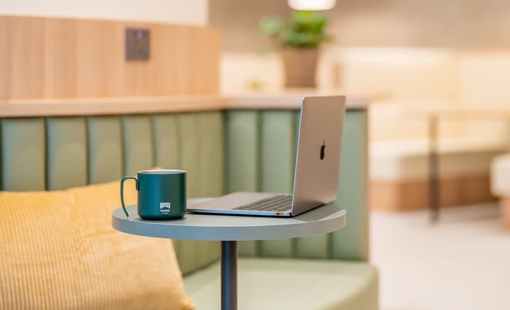 Close-up shot of an open laptop and a dark green mug resting on a small, round side table. The table is placed next to a curved, upholstered green couch in a brightly lit lounge area.