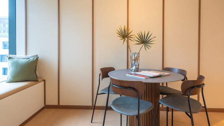 Detail shot of the modern dining area with a round grey table on a slatted wood base, four sleek wood and dark grey chairs, set against a panelled wall.
