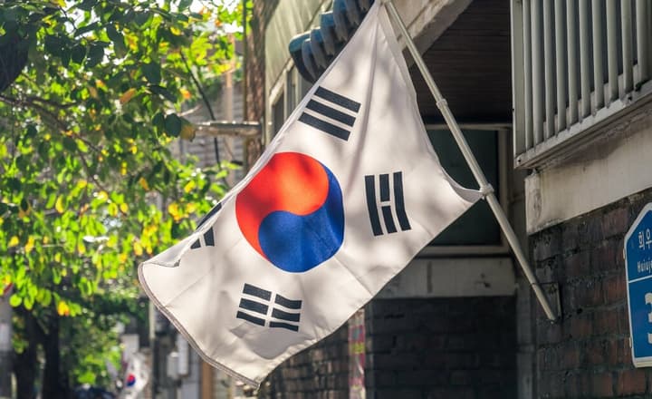 A South Korean flag (Taegeukgi) mounted on the side of a brick building, gently waving in the sunlight, with green tree leaves in the foreground.