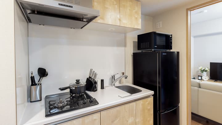Compact kitchen area with light wood-effect cabinets, a white counter, a black gas cooktop, a stainless steel sink, a black refrigerator, and a microwave on top in Monzennakacho.