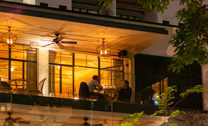 Night-time view of an outdoor restaurant terrace with a couple sitting at a table having a quiet dinner, illuminated by warm orange light from large grid-pane windows and hanging carriage-style lanterns.