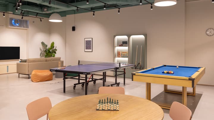 Bright recreational room featuring a ping pong table, a small billiards table, a round table with a chess set, and a distant sofa lounge area, all under a dark teal ceiling.