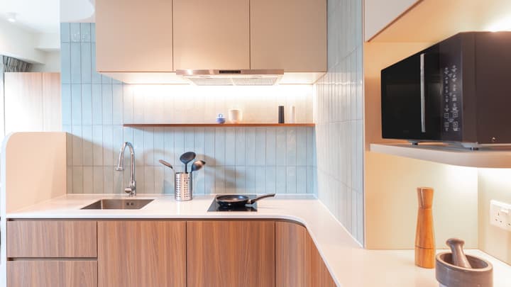 Close-up of a modern kitchenette featuring wood-grain lower cabinets, white counters, a sink, a two-burner cooktop, and a microwave on an open shelf, with a light blue ridged tile backsplash.