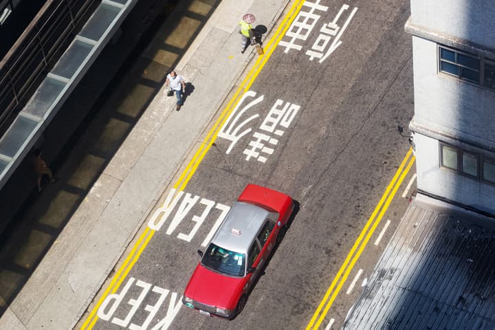 An aerial shot looking down onto a city street with a red and white taxi, yellow double lines, and Chinese characters painted on the asphalt.