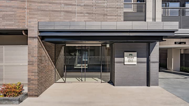 Exterior view of the main entrance to Weave Place Kunitachi apartment building, featuring a dark gray brick facade, a prominent black canopy structure, and glass double doors.